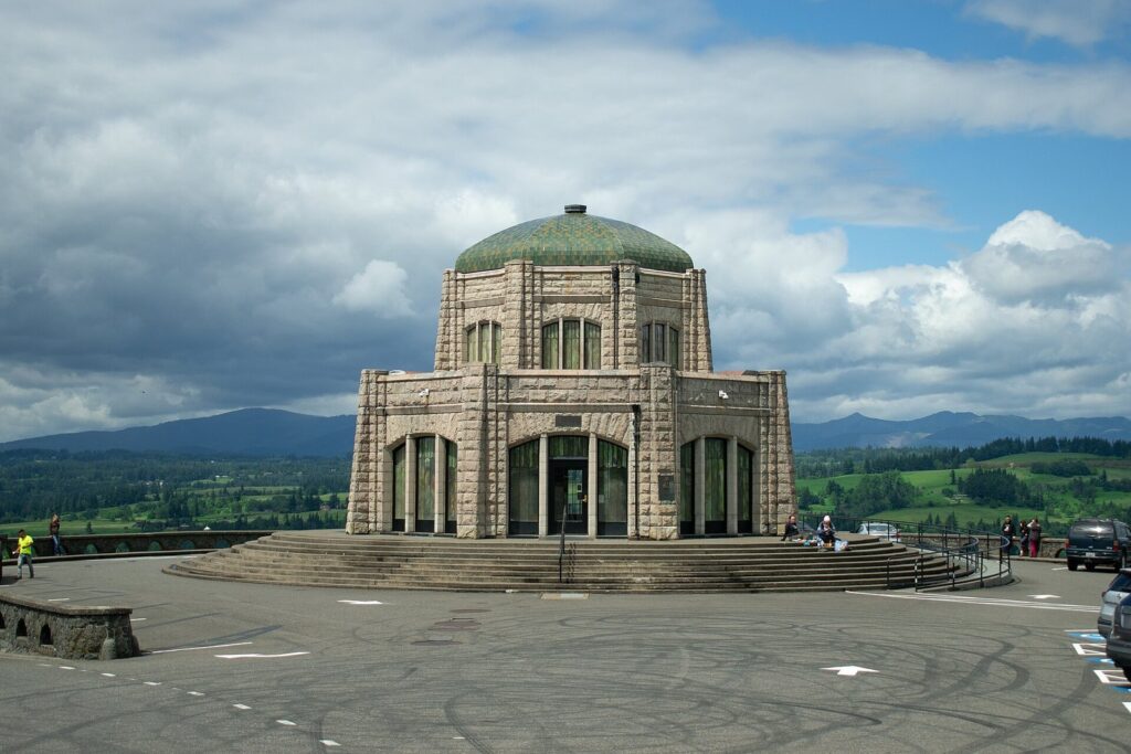 The domed stone Vista House on its round clifftop terrace under a partly cloudy sky, with green hills beyond