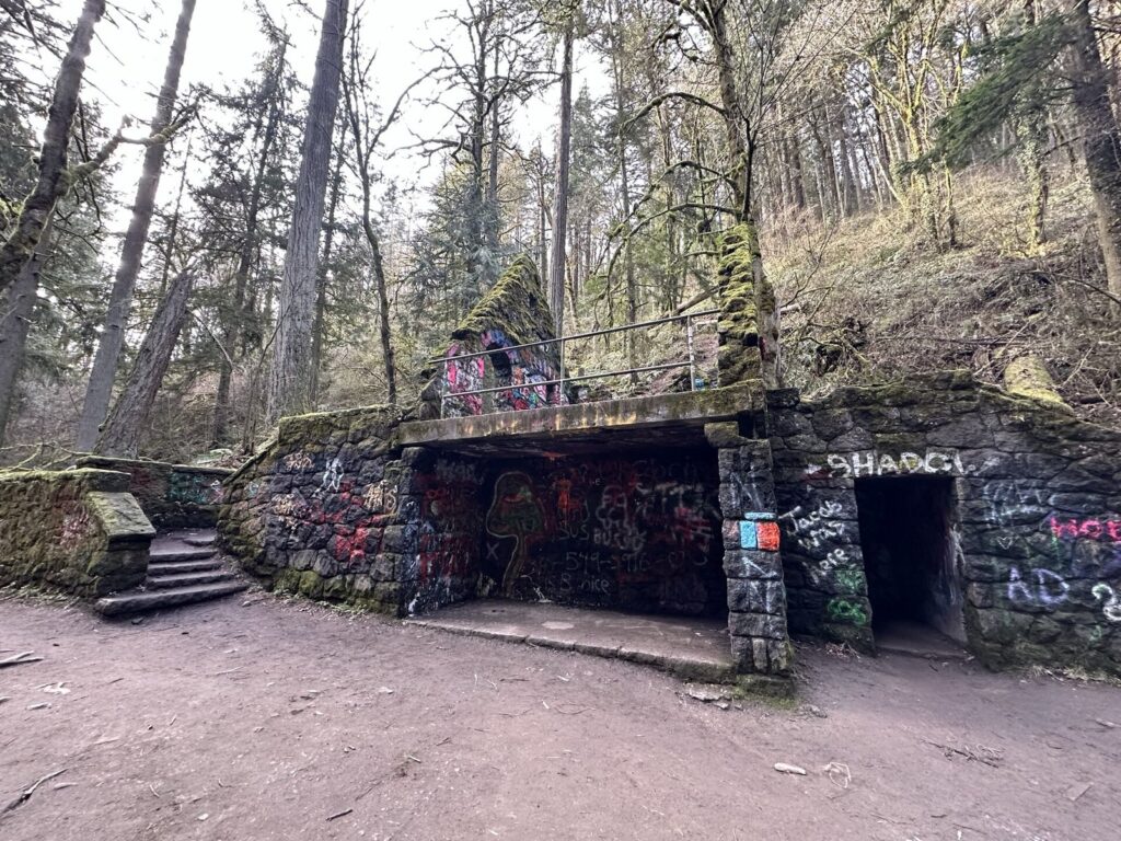 A roofless stone ruin covered in moss and graffiti beside a dirt trail in a forest of tall bare trees