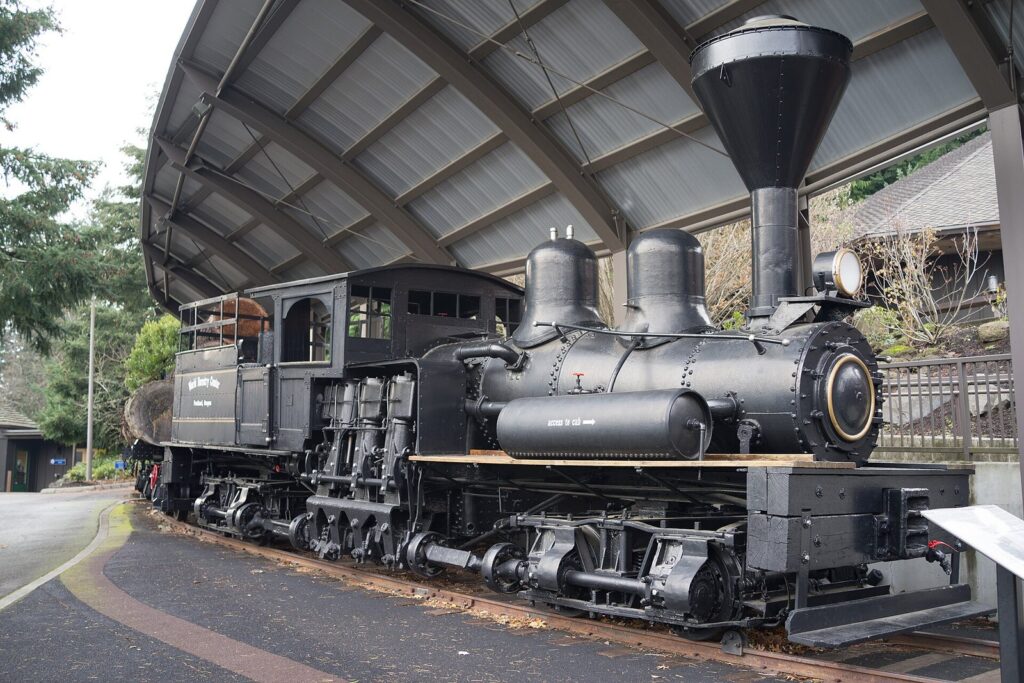 A black steam logging locomotive with a tall stack displayed under a curved metal canopy outside a museum